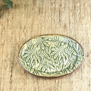 May include: An oval-shaped, ceramic serving dish with a raised floral pattern. The dish features a light green glaze with a darker brown rim. The floral design is a repeating pattern of stylized flowers and leaves.