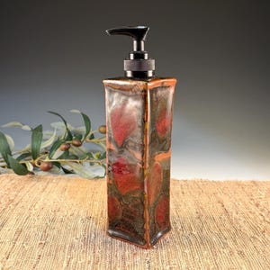 May include: A rectangular ceramic soap dispenser with a black pump. The dispenser has a mottled pattern of red, brown, and orange. The dispenser is on a woven surface with a sprig of leaves in the background.