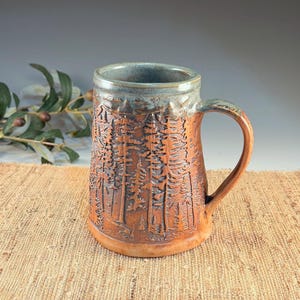 May include: A ceramic mug with a cylindrical body and a curved handle. The exterior features a textured, embossed forest scene with tall pine trees in a warm terracotta brown glaze, contrasted by a smooth, mottled blue-grey glaze at the rim.