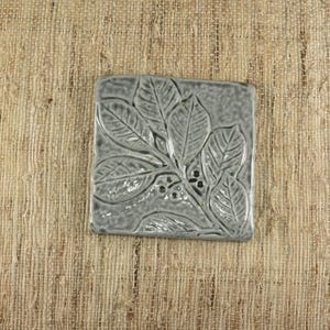 May include: A square, gray ceramic tile with a raised botanical design. The design features leaves and small berries. The tile is a muted gray color and has a slightly textured surface. The tile is likely used for home decor.