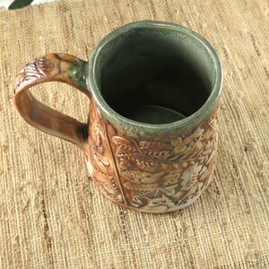 K&ouml;nnte beinhalten: Eine handgefertigte Keramik-Tasse mit einem detaillierten Reliefdesign aus Bl&auml;ttern und anderen Naturelementen. Die Tasse hat eine braune Au&szlig;enseite, einen gr&uuml;nen Rand und Innenraum. Der Griff ist ebenfalls braun und strukturiert.