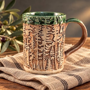 May include: A ceramic mug featuring a textured, carved forest scene with tall pine trees in shades of tan and brown. The rim and handle are glazed in a deep forest green, contrasting with the earthy, light-colored body of the vessel.