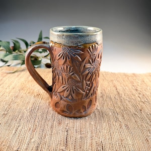 May include: A brown ceramic mug with a floral design. The mug has a handle and a blue-gray rim. The flowers are carved into the mug's surface. The mug is approximately 5 inches tall.