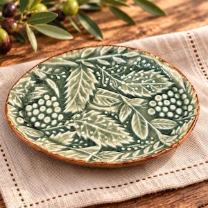 May include: A round ceramic plate featuring a raised, embossed pattern of green leaves and clusters of small white berries. The plate has a glossy finish with a dark green base color and lighter green highlights on the textured botanical design.