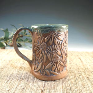 May include: A ceramic mug with a brown clay body featuring a relief pattern of coneflowers and leaves. The mug has a curved handle and a smooth, glossy green glaze applied to the upper rim, which drips slightly down onto the textured floral design.