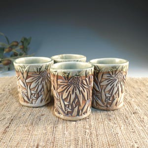 May include: Four ceramic shot glasses with a floral design. The glasses are tan with a carved sunflower pattern and a green rim. The glasses are approximately 3 inches tall.