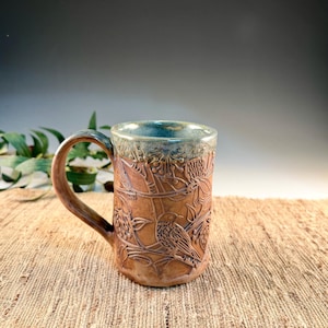May include: A handcrafted ceramic mug with a brown exterior and a blue-green interior. The mug features an embossed design of birds and foliage. The handle is brown, and the mug is set on a textured surface.