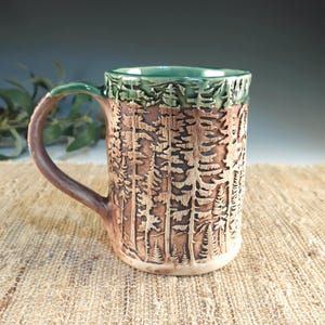 May include: A handmade ceramic mug with a forest design. The mug features a brown exterior with a carved tree pattern, a green rim, and a brown handle. The mug is approximately 4 inches in diameter and 5 inches tall.