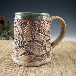 May include: A handcrafted ceramic mug with a detailed relief pattern of leaves and berries. The mug has a large handle and a wide opening. The rim is a muted green, and the body is a mix of tan and brown tones. The mug is approximately 6 inches tall.