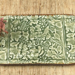 May include: A green ceramic serving platter with a detailed forest scene featuring animals and foliage. The oval-shaped platter has a brown rim and is decorated with embossed designs of foxes, owls, and rabbits. A small arrangement of dried flowers rests on the platter.