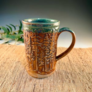 May include: A handmade ceramic mug with a forest scene carved into the exterior. The mug features a brown base, a tan body with a detailed tree design, and a teal-colored rim and handle. The mug is set on a textured, neutral-toned surface.