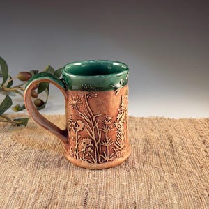 May include: A handmade ceramic mug with a brown exterior and a green interior and handle. The mug features an embossed floral design. The mug is sitting on a tan woven surface.