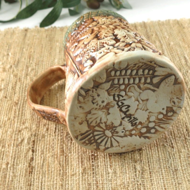 K&ouml;nnte beinhalten: Eine handgefertigte Keramik-Tasse mit einem detaillierten Reliefdesign aus Bl&auml;ttern und Laub. Die Tasse hat eine braune und beige Farbgebung mit einer strukturierten Oberfl&auml;che. Der Boden der Tasse zeigt eine Signatur und florale Muster.