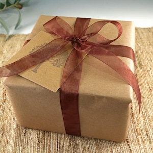 K&ouml;nnte beinhalten: Ein Geschenk in braunes Papier verpackt, mit einem transparenten, dunkelbraunen Band gebunden. Ein passendes Geschenkanh&auml;nger mit einem Baumdesign ist unter dem Band versteckt. Das Geschenk liegt auf einer strukturierten, beigefarbenen Oberfl&auml;che.
