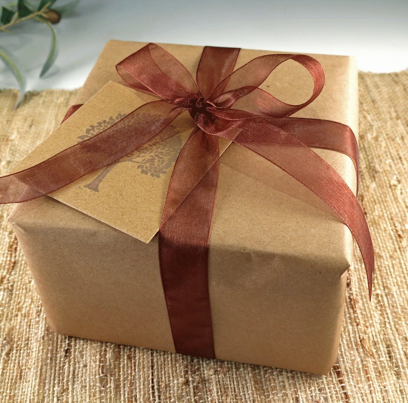 K&ouml;nnte beinhalten: Ein Geschenk in braunes Papier verpackt, mit einem transparenten, dunkelbraunen Band gebunden. Ein passendes Geschenkanh&auml;nger mit einem Baumdesign ist unter dem Band versteckt. Das Geschenk liegt auf einer strukturierten, beigefarbenen Oberfl&auml;che.