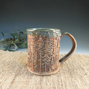 May include: A handmade ceramic mug with a forest design carved into the exterior. The mug features a brown and tan color scheme with a green rim and handle. The mug is sitting on a textured, tan surface.