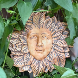 May include: A tan-colored ceramic wall plaque featuring a human face surrounded by a dense arrangement of sculpted leaves. The piece has dark, recessed detailing within the leaf veins and facial features, with the signature SEF etched on the side.