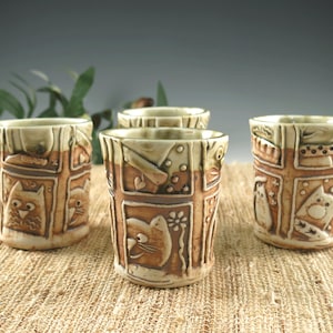 May include: Set of four ceramic mugs with a brown and green glaze. Each mug features a different design with a cat, bird, and flower motifs.