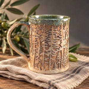 May include: A ceramic mug featuring a textured, carved forest scene with tall pine trees. The mug has a light tan base color with a dark, mottled green glaze around the rim and handle. The surface has a tactile, relief-like quality.