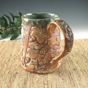 K&ouml;nnte beinhalten: Eine braune Keramiktasse mit einem detaillierten Reliefdesign aus Bl&auml;ttern, Eicheln und einer Eule. Der Griff und die Au&szlig;enseite sind strukturiert, mit einem gr&uuml;nen Rand. Die Tasse ist etwa 13 cm hoch.