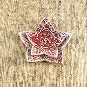 May include: A stack of three star-shaped ceramic ornaments. The ornaments are red with white snowflake and swirl designs. The ornaments are on a textured, tan surface.