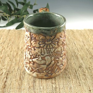 K&ouml;nnte beinhalten: Eine handgefertigte Keramiktasse mit einem detaillierten Reliefdesign von Waldtieren und Bl&auml;ttern. Die Tasse hat eine braune Au&szlig;enseite mit einem gr&uuml;nen Rand und Innenraum. Der Griff ist strukturiert, und die Gesamt&auml;sthetik ist rustikal und von der Natur inspiriert.