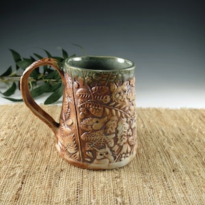 May include: A handmade ceramic mug with a nature-themed relief design. The mug has a brown exterior with detailed carvings of leaves, owls, and other forest elements. The handle is brown, and the rim has a green glaze.