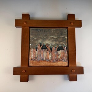 May include: A square ceramic tile featuring a relief carving of a forest with textured tree trunks and a dark, cloudy sky, set within a dark brown wooden frame with four protruding corners and small circular wooden pegs at each joint.