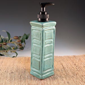 May include: A square, turquoise ceramic soap dispenser with a black pump. The dispenser has a textured, paneled design and a flared base. The background is a neutral color, with a sprig of olives on the left.