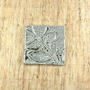 May include: Square, gray ceramic tile with a raised leaf and berry design. The tile has a glossy finish and is approximately 4 inches square. The leaves and berries are detailed, creating a textured surface. The tile is centered on a tan, woven background.