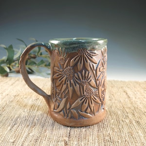 May include: A handmade ceramic mug with a brown base and a green rim. The mug features a detailed relief of sunflowers. The handle is textured, and the mug is approximately 5 inches tall.