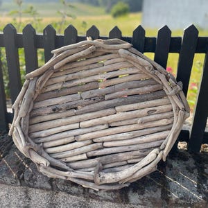 Könnte beinhalten: Ein achteckiges Tablett aus Treibholz. Es besteht aus zahlreichen kleinen, verwitterten Holzstücken, die eine natürliche, rustikale Ästhetik erzeugen. Die neutralen Farbtöne des Holzes passen zur Umgebung im Freien und deuten auf ein dekoratives oder Servierstück hin.