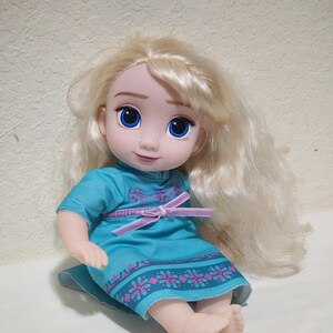 Puede incluir: Mu&ntilde;eca con pelo largo y rubio ondulado y ojos azules. La mu&ntilde;eca lleva un vestido turquesa con un dise&ntilde;o floral rosa y morado y un lazo rosa en la cintura. La mu&ntilde;eca est&aacute; sentada.