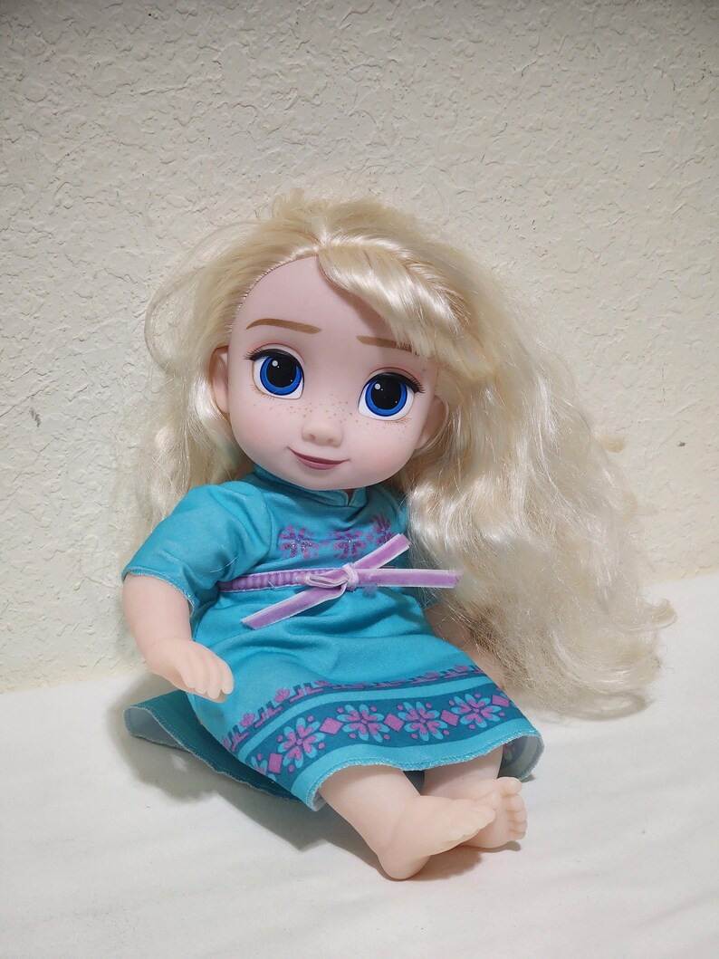 Puede incluir: Mu&ntilde;eca con pelo largo y rubio ondulado y ojos azules. La mu&ntilde;eca lleva un vestido turquesa con un dise&ntilde;o floral rosa y morado y un lazo rosa en la cintura. La mu&ntilde;eca est&aacute; sentada.