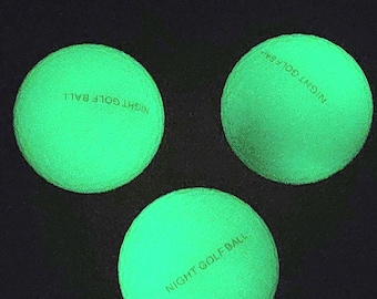 May include: Three glowing green golf balls against a black background. Each ball has the words "NIGHT GOLF BALL" printed on it. The balls are round and have a textured surface.