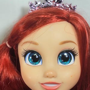 May include: A close-up of a doll's face with bright red hair and large blue eyes with glitter. The doll is wearing a pink tiara. The doll has a smiling expression and is wearing a purple dress.