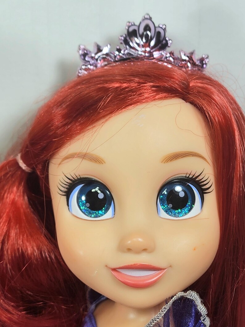 May include: A close-up of a doll's face with bright red hair and large blue eyes with glitter. The doll is wearing a pink tiara. The doll has a smiling expression and is wearing a purple dress.
