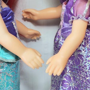 May include: Two dolls in princess-style dresses. One doll wears a teal and silver dress with a purple top. The other doll is in a purple dress with silver accents. Both dolls have their arms extended towards each other.