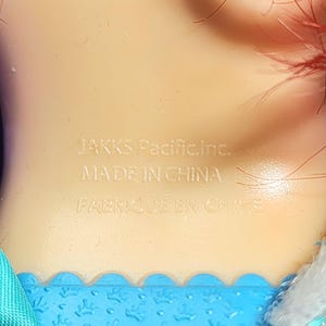 May include: Close-up of a doll's neck and shoulder area. The text "JAKKS Pacific, Inc. MADE IN CHINA FABRIQU&Eacute; EN CHINE" is imprinted on the doll's skin. Red hair is visible at the top of the frame, and blue fabric is at the bottom.