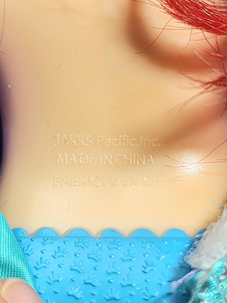 May include: Close-up of a doll's neck and shoulder area. The text "JAKKS Pacific, Inc. MADE IN CHINA FABRIQU&Eacute; EN CHINE" is imprinted on the doll's skin. Red hair is visible at the top of the frame, and blue fabric is at the bottom.
