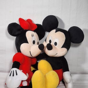 Pu&ograve; includere: Due personaggi Disney di peluche, Topolino e Minnie, sono mostrati vicini. Topolino indossa pantaloncini rossi, scarpe nere e guanti bianchi. Minnie indossa un vestito rosso, un fiocco rosso e guanti bianchi. Entrambi hanno orecchie nere e facce sorridenti.