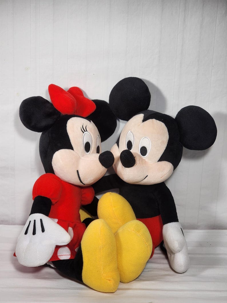 Pu&ograve; includere: Due personaggi Disney di peluche, Topolino e Minnie, sono mostrati vicini. Topolino indossa pantaloncini rossi, scarpe nere e guanti bianchi. Minnie indossa un vestito rosso, un fiocco rosso e guanti bianchi. Entrambi hanno orecchie nere e facce sorridenti.
