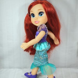 May include: A doll of Ariel, the Little Mermaid, with long red hair, blue eyes, and a purple tiara. She wears a purple and teal dress with matching purple shoes. The doll is in a walking pose.