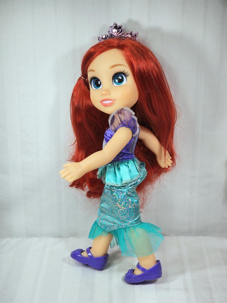 May include: A doll of Ariel, the Little Mermaid, with long red hair, blue eyes, and a purple tiara. She wears a purple and teal dress with matching purple shoes. The doll is in a walking pose.