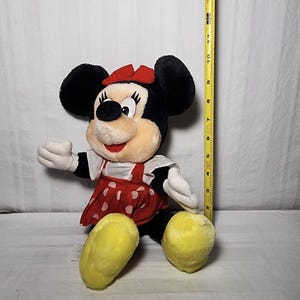 May include: A plush Minnie Mouse doll with black ears, a red bow, and a red dress with white polka dots. The doll has yellow feet and white gloves. A yellow measuring tape is next to the doll.
