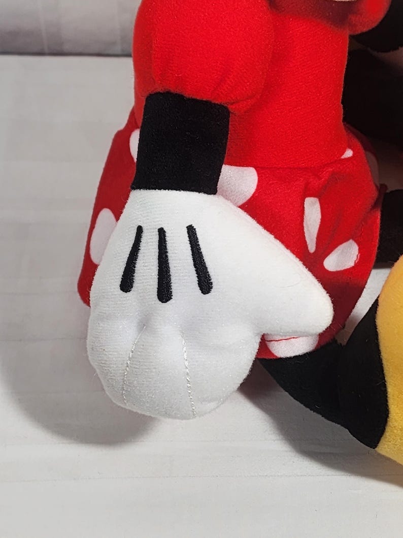 Pu&ograve; includere: Un peluche con una mano guantata bianca con linee nere, parte di un completo rosso a pois bianchi. Il giocattolo ha un polsino nero ed &egrave; visibile una scarpa gialla e nera.