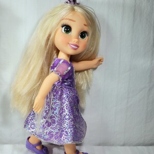 May include: A doll of a princess with long blonde hair, green eyes, and a tiara. She wears a purple dress with a floral pattern and matching purple shoes. The doll is posed as if walking.
