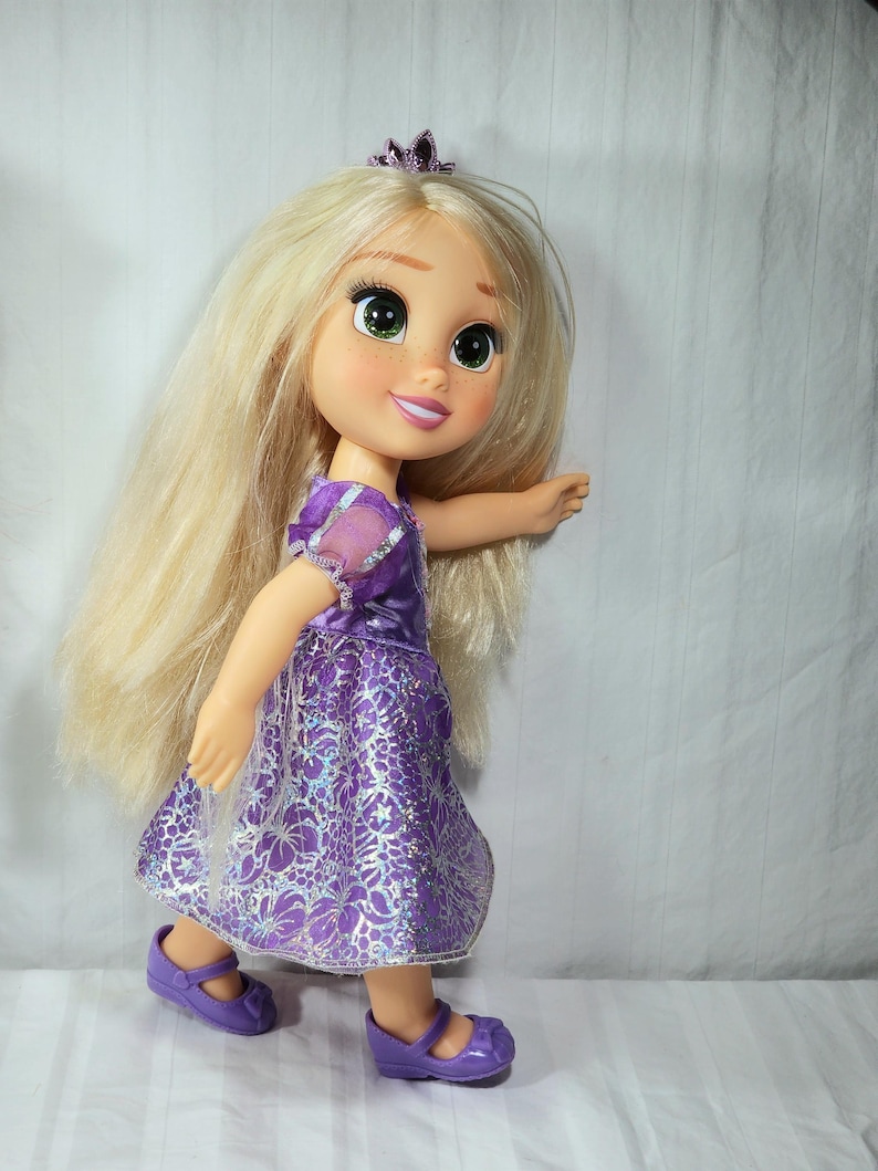 May include: A doll of a princess with long blonde hair, green eyes, and a tiara. She wears a purple dress with a floral pattern and matching purple shoes. The doll is posed as if walking.