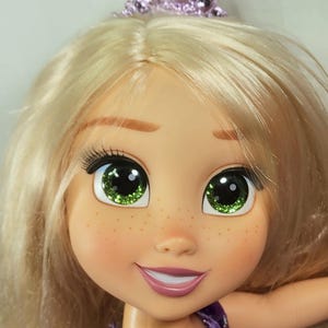 May include: A close-up of a doll's face with blonde hair and a purple tiara. The doll has green eyes with glitter, freckles, and a smiling expression. The doll is wearing a purple dress.