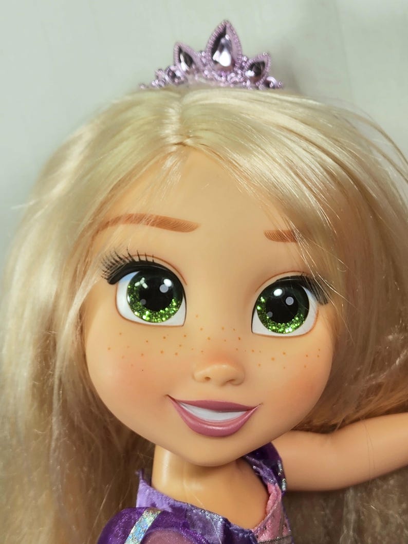 May include: A close-up of a doll's face with blonde hair and a purple tiara. The doll has green eyes with glitter, freckles, and a smiling expression. The doll is wearing a purple dress.